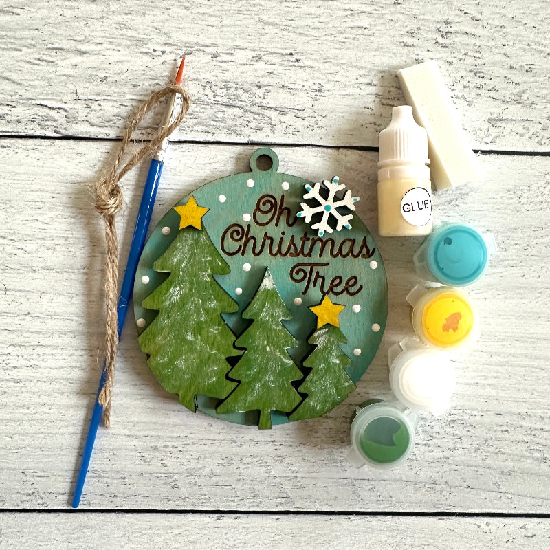 Christmas-themed wooden ornament with trees and text, surrounded by paint bottles and a brush on a wooden surface.