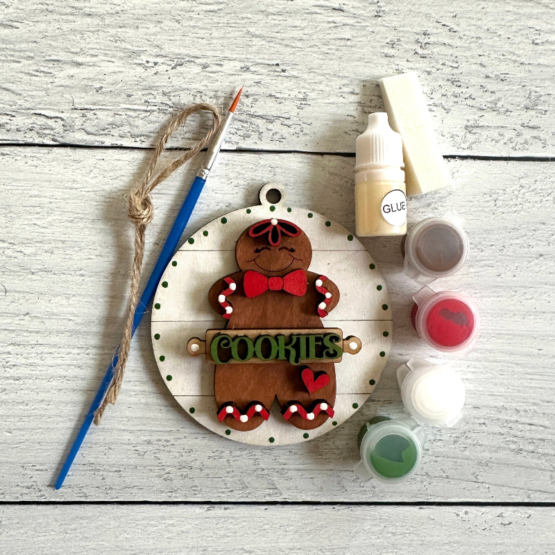 Decorative gingerbread man ornament with 'COOKIES' text on a wooden surface with paint supplies.